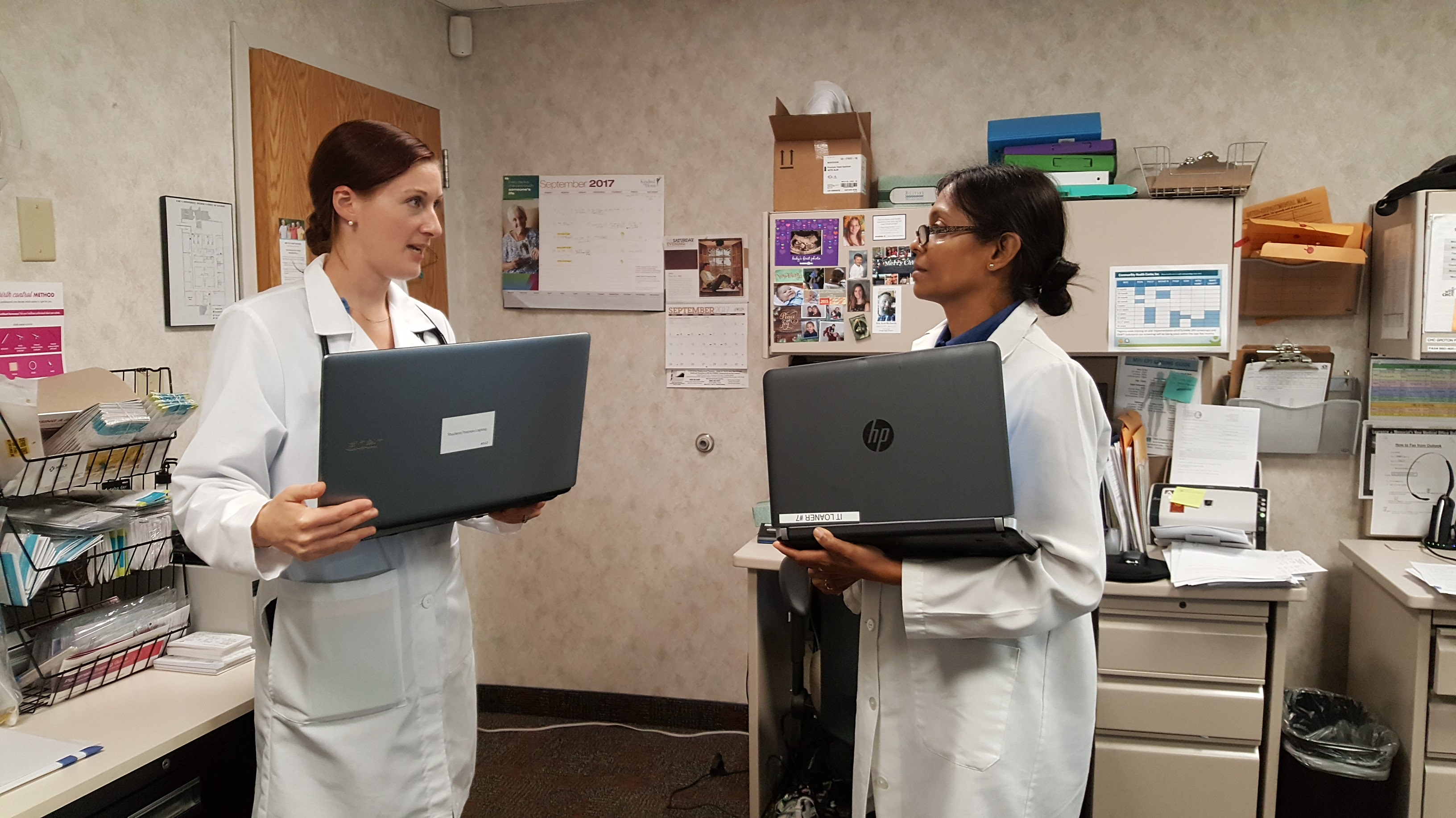 Two women in white coats talking to each other holding laptops in a doctors office